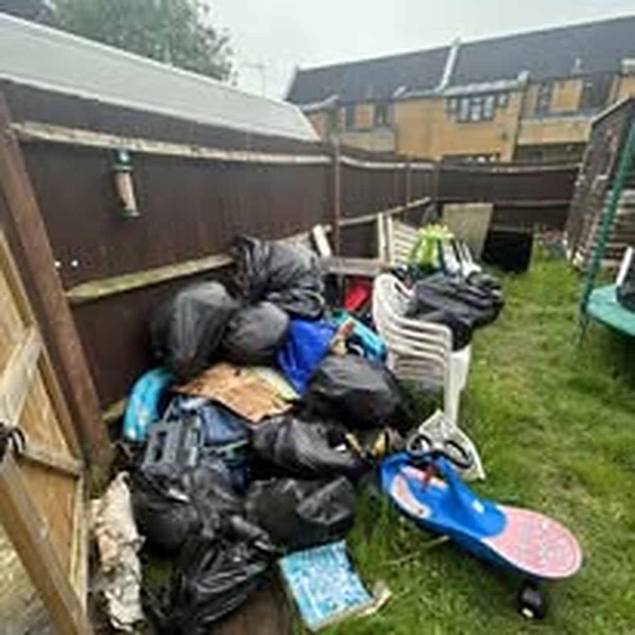 Garden piled high with black bags, boxes and household items against a wooden fence before clearance